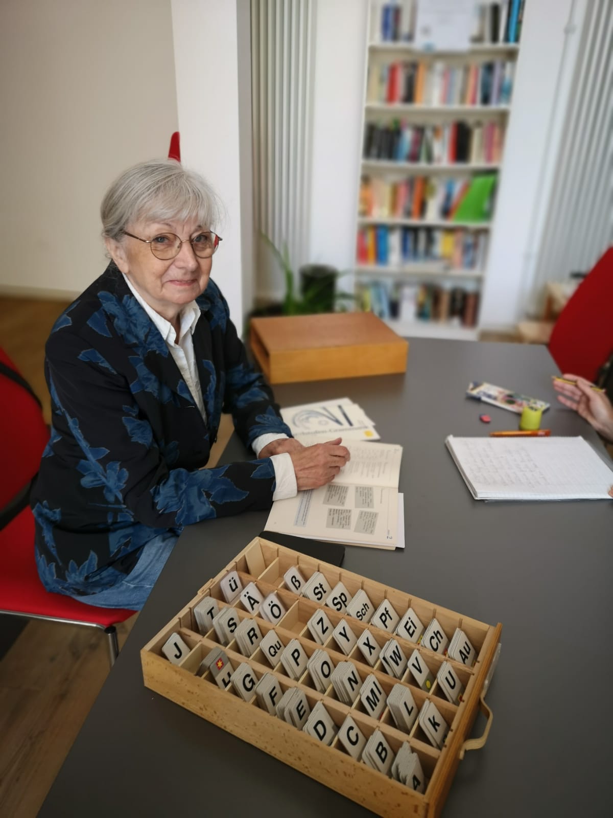 Frau mit grauen Haaren, Brille und blauem Blazer sitzt an einem Tisch, auf dem ein Kasten mit Buchstabenplatten steht
