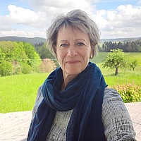Eine Frau mit kurzen, grauen Haaren lacht in die Kamera. Hinter ihr ist ein Feld mit Bäumen zu sehen.